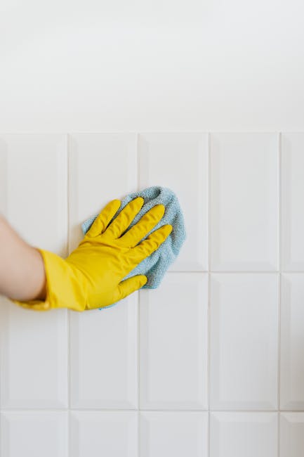 Close-up of a person wearing yellow rubber gloves holding a blue cleaning sponge in one hand and a clear spray bottle with a red cap in the other hand. The background shows a plain, light gray surface